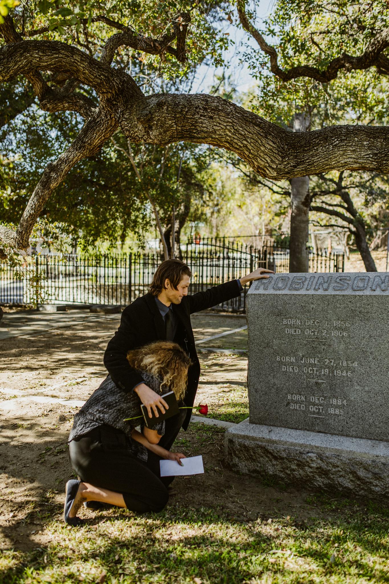 A couple mourning at a cemetery, embracing near a tombstone with a serene background.